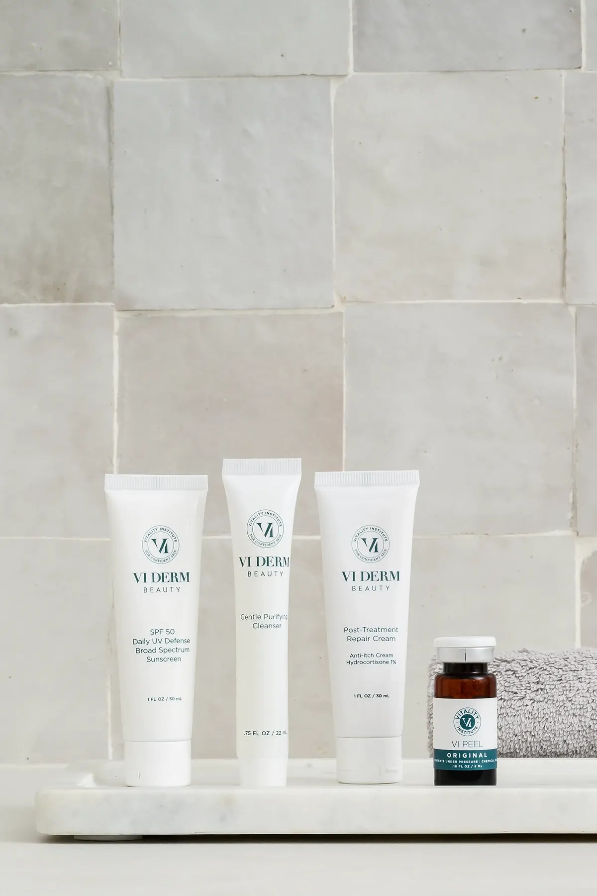 A minimalist display of VI Derm Beauty products, including SPF 50 Daily UV Defense Sunscreen, Gentle Purifying Cleanser, Post-Treatment Repair Cream, and VI Peel Original solution, arranged on a white marble tray with a folded gray towel, set against a tiled neutral background.