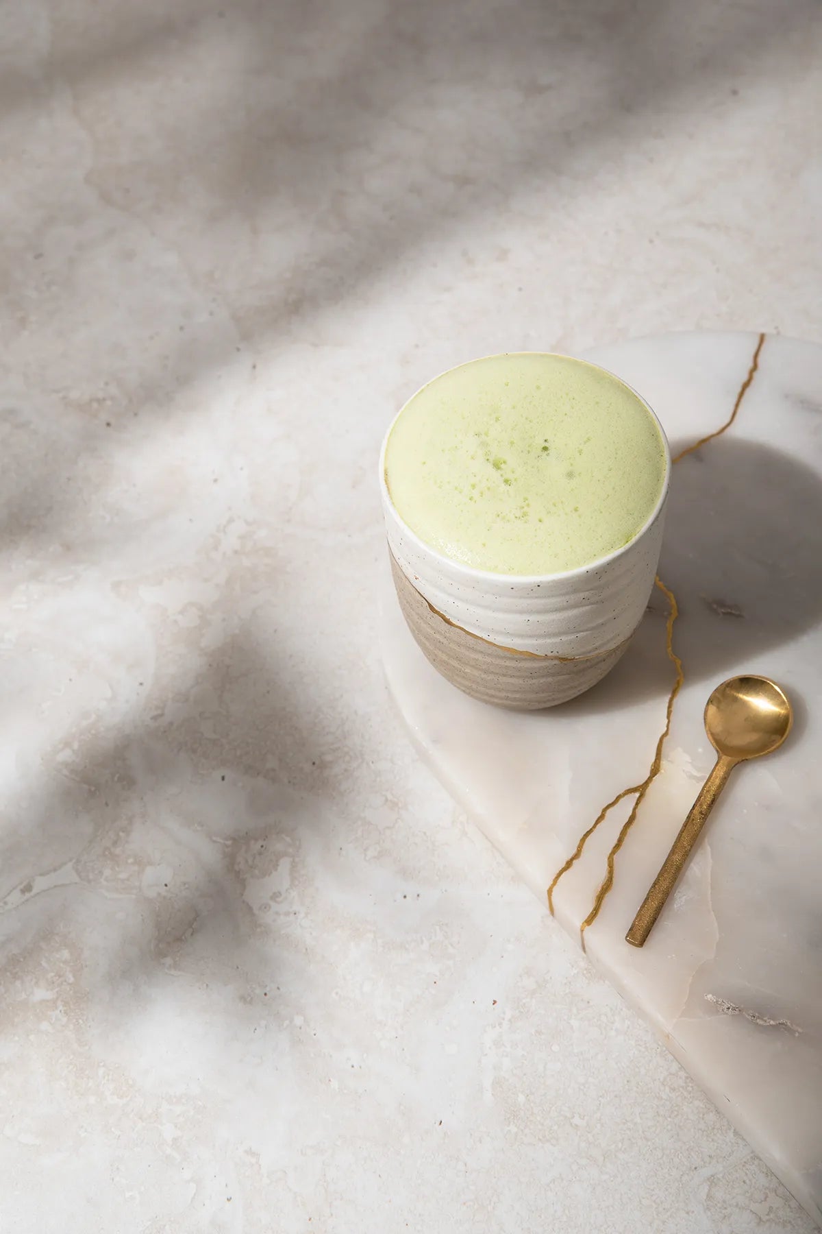 Matcha latte in a ceramic cup sitting on a white marble tray repaired with gold with gold spoon.